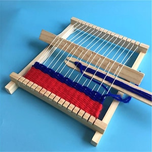 Op de afbeelding: Een houten weefgetouw met witte inslagdraden en een gedeeltelijk geweven textiel in rood en blauw. Het weefgetouw heeft een rechthoekig frame met inkepingen voor de inslagdraden en een houten schietspoel.