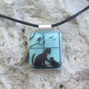 May include: A square pendant necklace featuring a black silhouette of two cats looking out a window with birds and a tree branch. The pendant has a light blue background and is attached to a black cord.