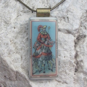 Può includere: Una collana con pendente rettangolare con un'illustrazione vintage di una figura a cavallo, con testo in francese. Il pendente ha un'ansa color ottone e pende da una sottile catena dorata.