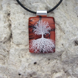 Puede incluir: Un collar con un colgante de vidrio y un cordón de cuero negro. El colgante presenta un árbol blanco con raíces sobre un fondo rojo y negro.