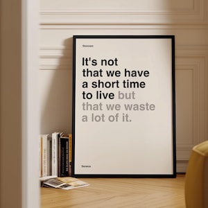 May include: A framed black and white print with the quote "It's not that we have a short time to live but that we waste a lot of it." The print is propped against a wall, with a stack of books and a yellow ottoman in the background.