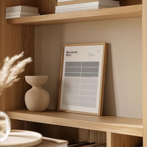 May include: A framed print with the words "Memento Mori" in a minimalist design. The print features a grid pattern with black and white squares. The frame is a light brown color, and the print is displayed on a wooden shelf.