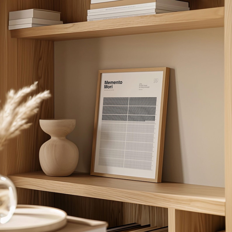 May include: A framed print with the words "Memento Mori" in a minimalist design. The print features a grid pattern with black and white squares. The frame is a light brown color, and the print is displayed on a wooden shelf.