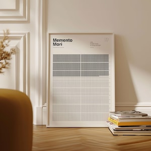 May include: A framed art print with the words "Memento Mori" in bold black font. The print features a grid-like design with black and white squares. A stack of books rests on the wooden floor in front of the print.