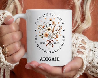 May include: A white ceramic mug featuring a printed bouquet of wildflowers in shades of orange, brown, and cream. The text reads "CONSIDER HOW THE WILDFLOWERS GROW" in a circular arc, "LUKE 12:27" in the center, and the name "ABIGIAL" at the bottom.