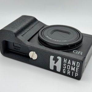Può includere: Fotocamera nera con obiettivo e impugnatura protettiva. L'impugnatura presenta la scritta bianca "HANDSOME GRIP". L'obiettivo della fotocamera riporta la scritta "GR LENS f=18.3mm 1:2.8".