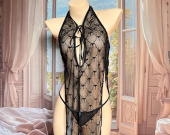 Könnte beinhalten: Ein schwarzes, transparentes Spitzen-Neckholder-Kleidungsstück mit einem sich wiederholenden Muster aus kleinen kreisförmigen Motiven. Das Teil hat einen tiefen V-Ausschnitt mit schwarzer Schnürung in der Mitte und einen passenden schwarzen Tanga.