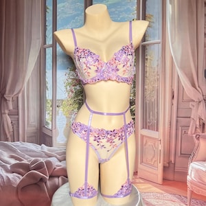 May include: A lavender lingerie set featuring a sheer bra and matching high-cut panties with intricate floral embroidery in shades of purple and pink. The set includes a waist strap and two leg garters, all connected by thin lavender satin straps.