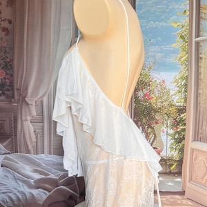 K&ouml;nnte beinhalten: Ein wei&szlig;es &auml;rmelloses Kleidungsst&uuml;ck mit einem ger&uuml;schten Oberteil und einem transparenten Spitzenrock. Das Oberteil hat d&uuml;nne Spaghettitr&auml;ger und ein r&uuml;ckenfreies Design, w&auml;hrend der untere Teil ein florales Spitzenmuster auf wei&szlig;em Futter zeigt.