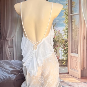 K&ouml;nnte beinhalten: Ein wei&szlig;es Slip-Kleid mit tiefem R&uuml;ckenausschnitt und d&uuml;nnen Spaghettitr&auml;gern, die sich in der Mitte kreuzen. Der obere R&uuml;ckenbereich ist mit geschichteten R&uuml;schen verziert, w&auml;hrend der untere Teil des Kleides aus transparenter wei&szlig;er Spitze besteht.