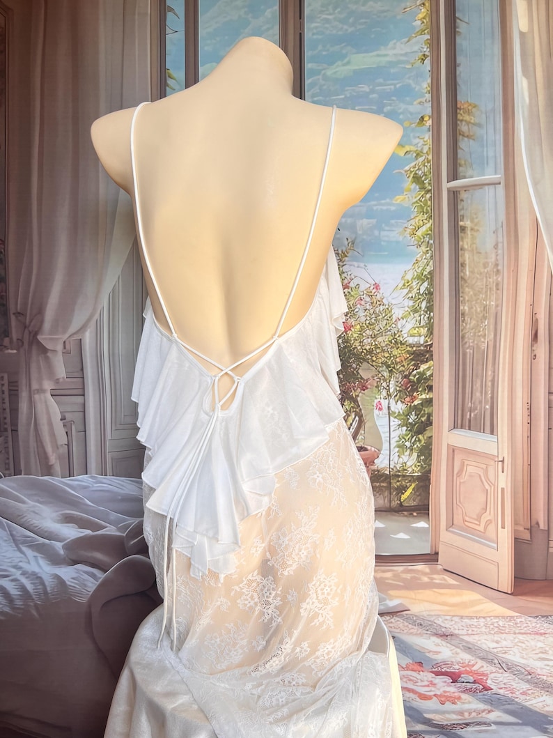 K&ouml;nnte beinhalten: Ein wei&szlig;es Slip-Kleid mit tiefem R&uuml;ckenausschnitt und d&uuml;nnen Spaghettitr&auml;gern, die sich in der Mitte kreuzen. Der obere R&uuml;ckenbereich ist mit geschichteten R&uuml;schen verziert, w&auml;hrend der untere Teil des Kleides aus transparenter wei&szlig;er Spitze besteht.