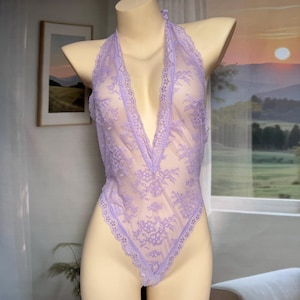 May include: A lavender lace bodysuit with a plunging neckline and halter straps. The lingerie features floral lace detailing and a high-cut leg. The garment is displayed on a mannequin.