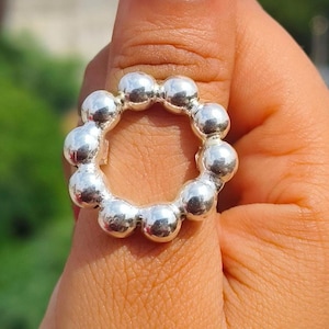 May include: A silver ring featuring a circular design composed of small, evenly spaced spheres. The ring is shown on a finger, with a blurred green background. The ring's metallic surface reflects light, creating highlights.