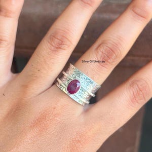May include: A silver-colored ring with an oval, deep red gemstone. The ring has a detailed, etched design around the band. The ring is on a finger. The text "SilverGiftArtisan" is visible.