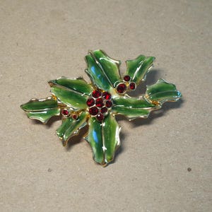 May include: A gold-tone metal brooch shaped like holly leaves and berries. The leaves are finished with green enamel, and the clusters of berries are set with small, round, red faceted crystals that catch the light with a brilliant sparkle.