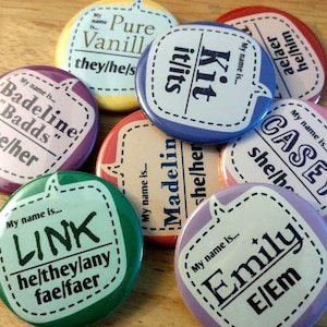 Könnte beinhalten: Eine Sammlung bunter, runder Buttons mit verschiedenen Namen und Pronomen. Jeder Button hat ein Sprechblasendesign mit Namen und Pronomen wie "she/her" oder "they/he/s". Die Buttons liegen auf einer Holzoberfläche.