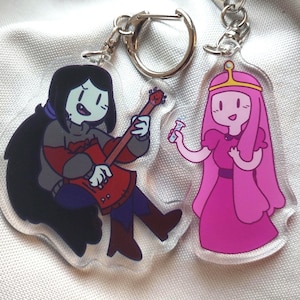 May include: Two clear acrylic keychains featuring cartoon characters. One keychain depicts a character with black hair playing a red guitar. The other keychain shows a character in a pink dress and crown holding a small vial.