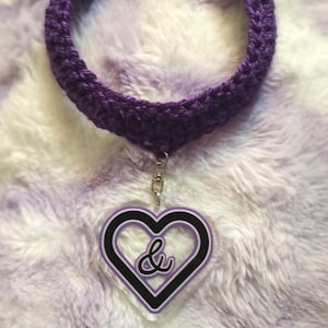 May include: A purple crochet choker necklace with a silver clasp. The necklace features a heart-shaped pendant with a black outline and a white ampersand symbol. The background is a soft, fluffy, purple and white fabric.
