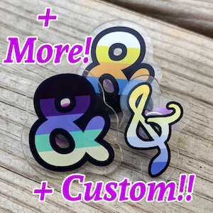 May include: Three clear acrylic pins shaped like ampersands, each featuring horizontal stripes in different color combinations including yellow, orange, blue, purple, teal, and light green. The text "+ More!" and "+ Custom!!" appears in purple.