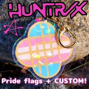 May include: A clear, four-leaf clover-shaped keychain with a gold clasp. The keychain features a design with pink, blue, and white colors, and the text "HUNTRAX" and "Pride flags + CUSTOM!" in pink.