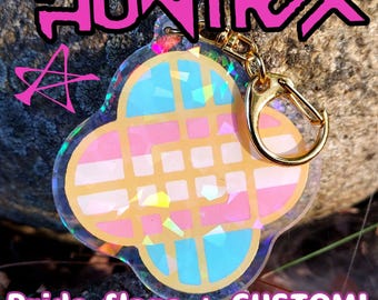 May include: A clear, four-leaf clover-shaped keychain with a gold clasp. The keychain features a design with pink, blue, and white colors, and the text "HUNTRAX" and "Pride flags + CUSTOM!" in pink.