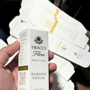 Puede incluir: Una caja blanca de aceite facial Swacci Flora Radiance, 30 ml. La caja presenta un diseño floral y el texto "Nature's Luxury". Los ingredientes están listados en el lateral. La caja se sostiene en una mano, con otras cajas en el fondo.