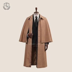 Può includere: Un cappotto a mantella in lana color marrone chiaro indossato sopra una giacca da completo nera gessata, una camicia bianca con colletto e una cravatta nera. Il cappotto presenta un colletto strutturato, chiusura con bottoni e mantella integrata. Il testo Bespoke Atelier London è visibile.