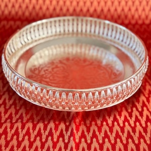 May include: A round, silver-colored tray with an ornate, openwork border. The tray's interior has a subtle, embossed pattern. The tray is set against a vibrant red background with a repeating geometric design, creating a striking contrast.