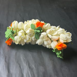 May include: A floral hair accessory featuring a dense arrangement of cream-colored jasmine-like blossoms interspersed with vibrant orange petals and small green leaves, creating a rounded, garland-like structure for hair styling.