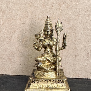 May include: A small, gold-colored statue of a seated deity. The figure wears an ornate headdress and holds objects in its hands. The statue is on a rectangular base with detailed carvings. The background is a neutral tan color.