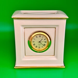 May include: A cream-colored mantel clock with gold trim and a round clock face. The clock face has Roman numerals and gold hands. The clock is rectangular with a small opening at the top and sits against a green background.