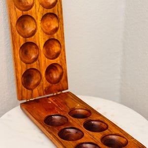 May include: A rectangular wooden board game with a hinged design, featuring two rows of four circular indentations on each side. The board is crafted from polished brown wood with a visible grain and includes metal hinges and a small metal latch.