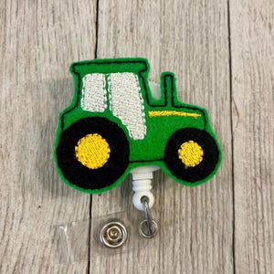 May include: Green tractor badge reel with black and yellow wheels, white window, and yellow accents. The badge reel has a clear plastic strap and a metal clip for attaching to clothing. The tractor is an embroidered design.