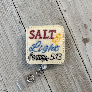 May include: A square, off-white badge reel with the words "SALT & LIGHT Matthew 5:13" embroidered in burgundy, blue, yellow, and black thread. The badge reel has a clear plastic strap and a white, round clip.