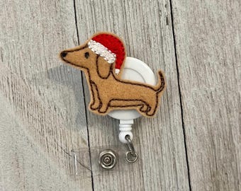May include: A brown dachshund dog wearing a red and white Santa hat, attached to a white retractable badge reel. The badge reel has a clear plastic strap and a metal snap closure. The background is a light wood grain.