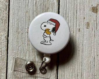 Può includere: Un portabadge circolare bianco con l'illustrazione del personaggio dei cartoni animati Snoopy che indossa un cappello da Babbo Natale rosso e tiene in mano un piccolo uccellino giallo. Include un cinturino in plastica trasparente con bottone.