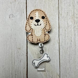 Puede incluir: Un carrete de insignia de perro bordado en beige y blanco con un dije de hueso. El perro tiene ojos y nariz negros, con costuras marrones en los bordes. El dije de hueso es blanco con costuras negras.