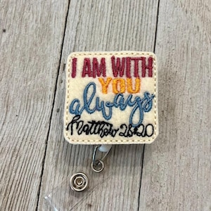 Könnte beinhalten: Ein quadratisches, cremefarbenes Ausweis-Jojo mit dem gestickten Text "I AM WITH YOU always Matthew 28:20" in Burgunderrot, Gelb und Blau. Das Ausweis-Jojo hat ein einziehbares Kabel und einen Metallclip.