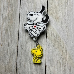 Può includere: Un porta badge di Snoopy bianco e nero con un cuore rosso e un ciondolo Woodstock giallo. Il design di Snoopy è in posa rilassata. Il porta badge è attaccato a un cordino bianco.