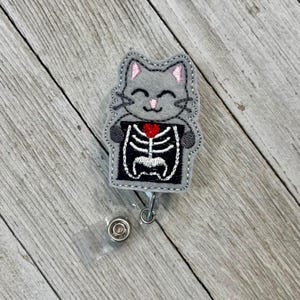 Puede incluir: Un portatarjetas con forma de gato gris con una cara sonriente y detalles en rosa. El gato sostiene una imagen de rayos X en blanco y negro con un corazón rojo. El portatarjetas tiene una correa de plástico transparente y un clip de metal.