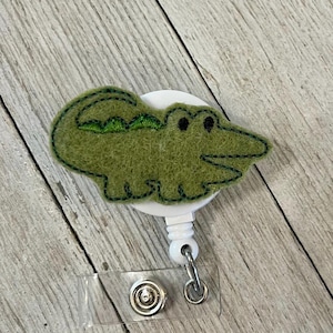 May include: A green felt alligator badge reel with black stitched details. The alligator is attached to a white circular base, with a retractable reel and a clear plastic strap with a metal snap.