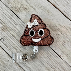 May include: A brown glitter badge reel featuring a smiling poop emoji with a white bow. The emoji has black eyes and a white embroidered smile. The reel has a clear plastic strap and a metal clip.