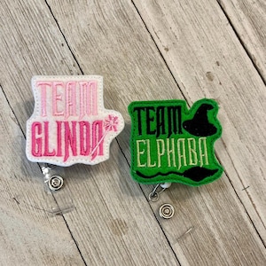 Könnte beinhalten: Zwei Filz-Ausweisrollen. Eine ist rosa und weiß mit dem Text "TEAM GLINDA" und einem Sternmuster. Die andere ist grün mit dem Text "TEAM ELPHABA" und einem Hexenhut- und Schnurrbart-Design.