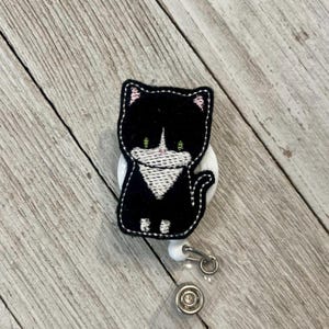 Puede incluir: Un portatarjetas de gato bordado en blanco y negro. El gato tiene orejas rosas y ojos verdes. El portatarjetas tiene una base blanca y un cordón retráctil con un clip metálico.