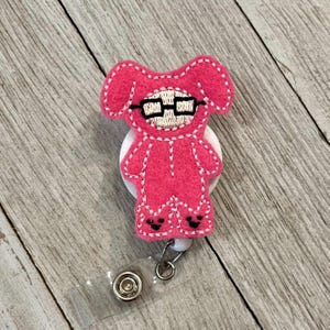 Pu&ograve; includere: Un porta badge in feltro rosa con un orsetto dei cartoni animati con gli occhiali. L'orso &egrave; cucito con filo bianco e ha accenti neri sui piedi. Il porta badge ha un cinturino in plastica trasparente e una clip in metallo.