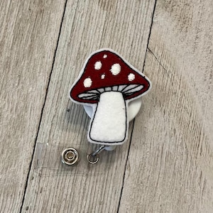 May include: A red and white mushroom-shaped badge reel with a black outline. The mushroom cap has white spots. The badge reel has a clear plastic strap and a metal snap closure.