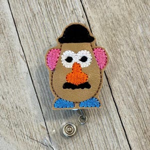 May include: A tan badge reel depicting a cartoon potato character with a black hat, white eyes, pink ears, an orange nose, a black moustache, and blue shoes. The badge reel has a clear plastic strap and a metal clip.