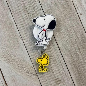 Op de afbeelding: Een wit en zwart Snoopy badge reel met een rode kraag en een gele Woodstock bedel. Het Snoopy-ontwerp is bevestigd aan een witte oprolhaspel. De Woodstock bedel hangt eronder.