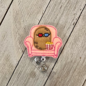 Puede incluir: Una insignia de fieltro con un personaje de patata con gafas 3D sentado en un sofá rosa. Un pequeño recipiente de palomitas de maíz está al lado de la patata. La insignia tiene un carrete retráctil y una correa de plástico transparente.