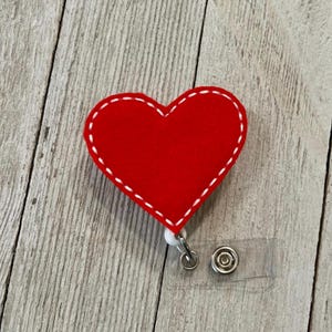 May include: A red heart-shaped badge reel with white stitching around the edges. The badge reel has a clear plastic strap and a metal clip for attaching to clothing. The background is a light wood grain.